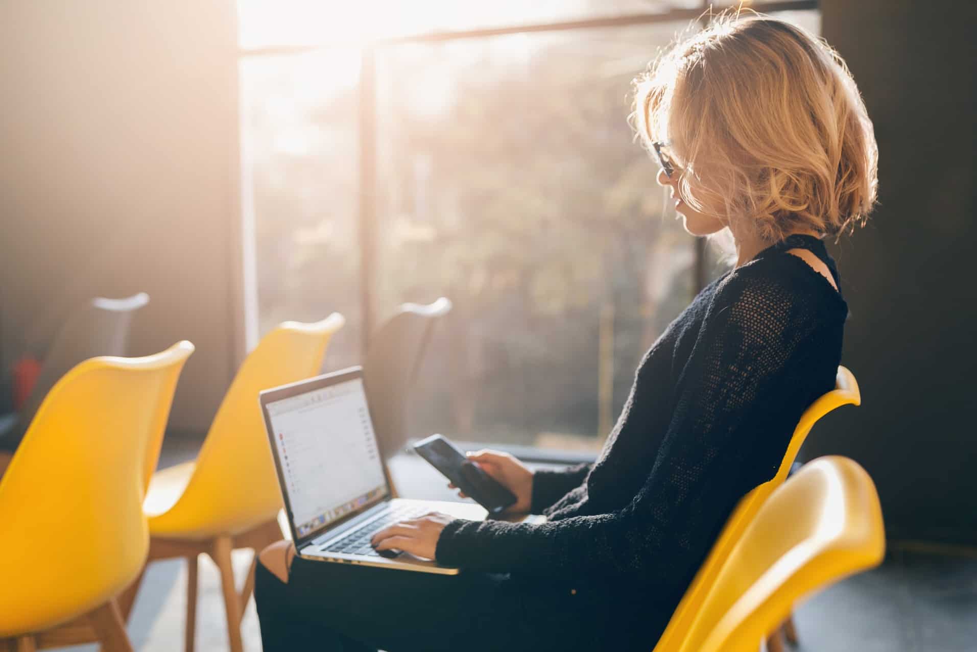 Woman sitting in a yellow chair looking at a laptop and her phone