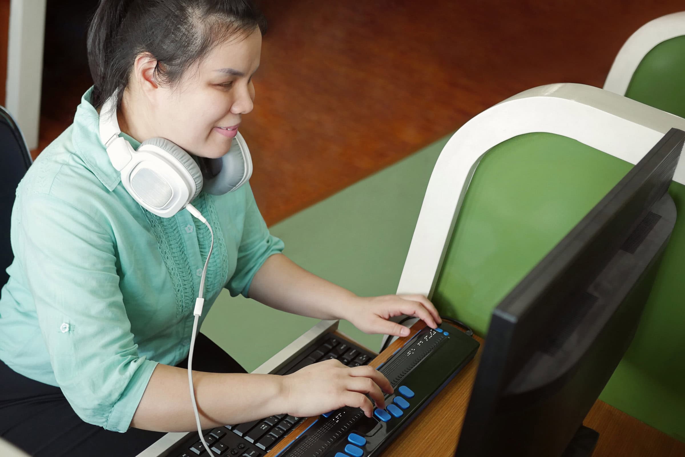 asian young blind woman with headphones using computer with refreshable braille display or braille terminal a technology device for persons with visual disabilities.